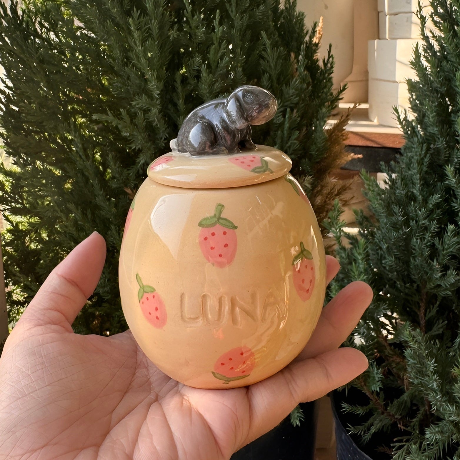 A hand holds a small beige ceramic jar with a lid, decorated with pink strawberries and the name 'LUNA'. A small gray figurine sits on top of the lid.